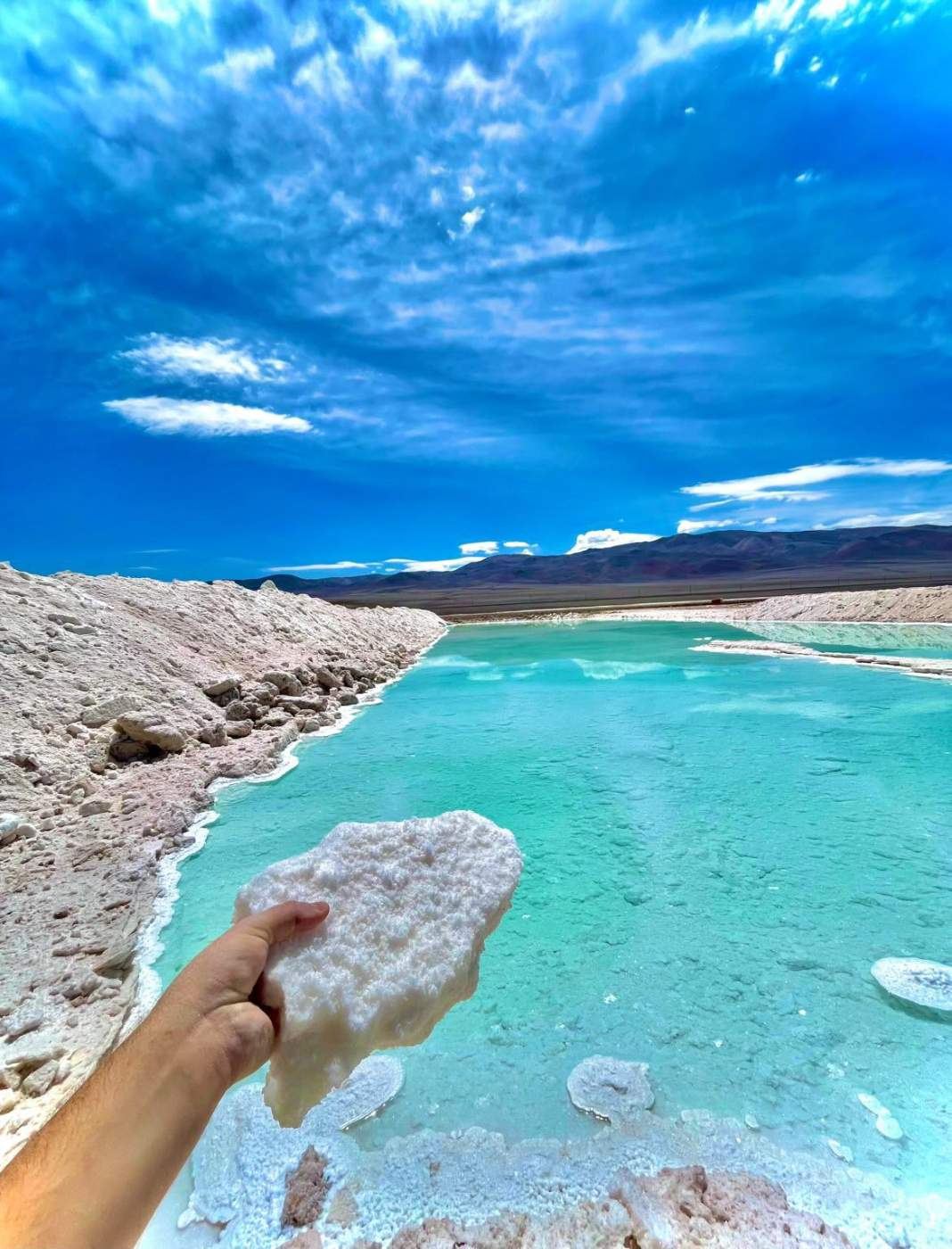 Mano sosteniendo un trozo de sal frente a una laguna turquesa del salar